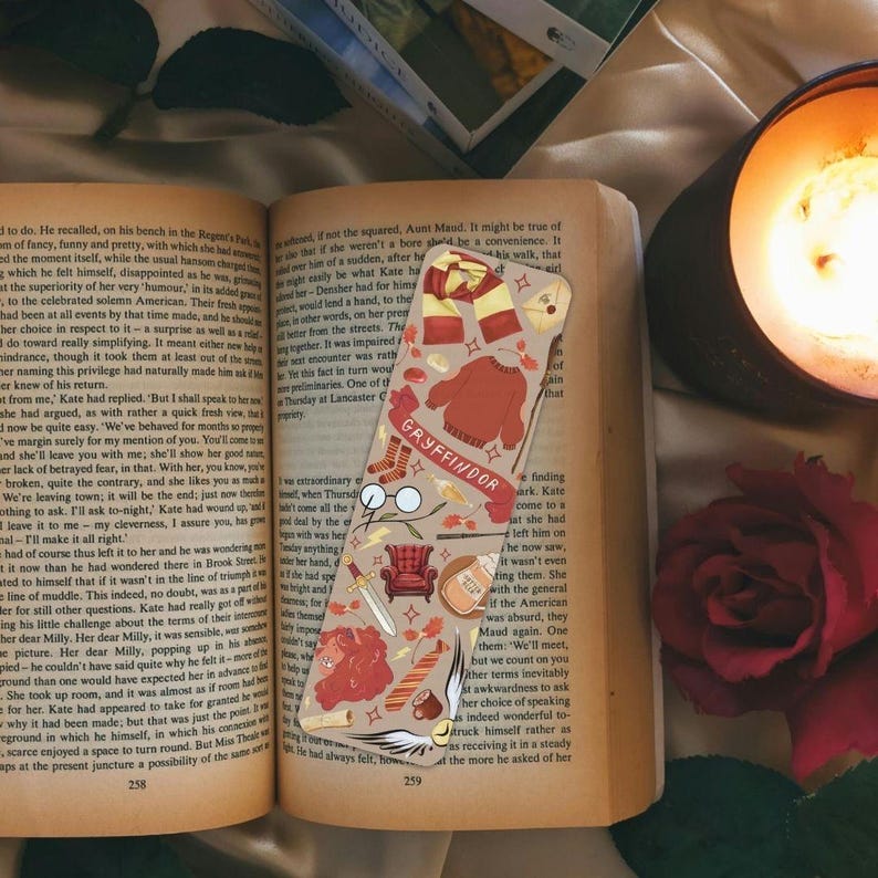 Op de afbeelding: Een Gryffindor boekenlegger met illustraties van een trui, sjaal en laarzen in rood, goud en cr&egrave;me. De boekenlegger is geplaatst in een open boek, met het woord 'Gryffindor' zichtbaar.