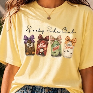 Op de afbeelding: Een lichtgeel t-shirt met de tekst Spooky Soda Club boven vier frisdrankblikjes met strikken. De blikjes zijn gelabeld als Root Fear met pompoenen, Dr Ghoul met een vampier, Mtn Boo met vleermuizen en een kerkhof, en Diet Spook met een geest.