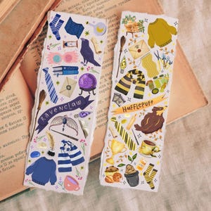 Op de afbeelding: Twee decoratieve bladwijzers met illustraties. E&eacute;n toont een raaf, een stropdas en de tekst "Ravenclaw". De andere toont een das, een sjaal en de tekst "Hufflepuff". Beide hebben een vintage uitstraling.