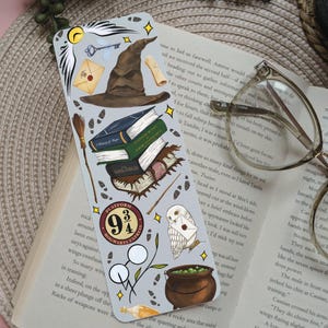 May include: A bookmark with illustrations of a sorting hat, books, a key, an owl, and a cauldron. The bookmark has a light grey background with yellow star accents and the text "Platform 9 3/4".