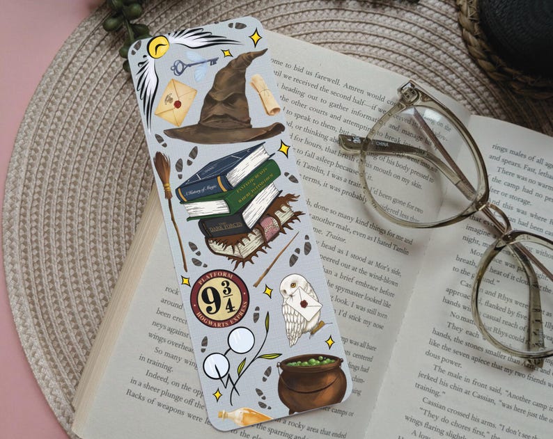 May include: A bookmark with illustrations of a sorting hat, books, a key, an owl, and a cauldron. The bookmark has a light grey background with yellow star accents and the text "Platform 9 3/4".