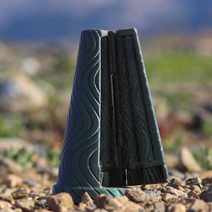 Puede incluir: Un objeto en forma de cono de color verde oscuro con un diseño de vetas de madera texturizadas. El objeto está colocado sobre una superficie rocosa con un fondo borroso de montañas y cielo azul. El objeto parece ser un marcador o una pieza decorativa.