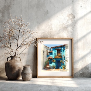 May include: A framed watercolour print depicting a vivid blue building with a red-tiled roof and balcony, set against a clear blue sky. The print is in a light wooden frame, next to a vase with branches and a smaller vase.
