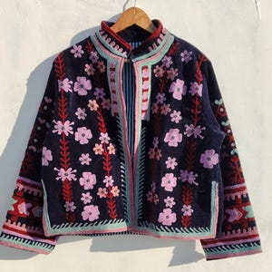 May include: A navy blue jacket with floral embroidery. The jacket features pink, red, and teal accents, with a high collar and a full-length zip. The floral design is spread across the front and sleeves.