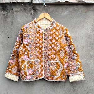 May include: A quilted jacket with a floral pattern in shades of gold, pink, and blue. The jacket has a round collar, two front pockets, and is lined with a striped fabric. The jacket is hanging on a wooden hanger.
