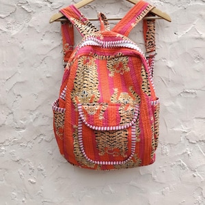 May include: A quilted backpack with a vibrant tiger stripe pattern in shades of orange, pink, and gold. The backpack has adjustable straps, a front pocket, and side pockets. It is hanging on a wooden hanger against a textured white wall.