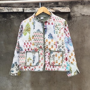 May include: White quilted jacket with a colorful floral pattern. The jacket features a mandarin collar, two front pockets, and a black and white striped trim. The jacket is hanging on a wooden hanger.