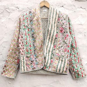 Peut inclure: Veste matelassée avec un motif patchwork. Elle présente des panneaux floraux, rayés et unis dans des tons de vert, rose et beige. La veste est suspendue à un cintre en bois sur un mur blanc texturé.
