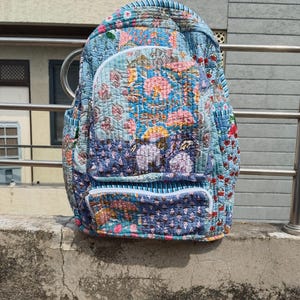 May include: A patchwork backpack with a floral design in blue, pink, and yellow. The bag has a front pocket with a white zip and two side pockets. The straps and top handle are visible. The bag is made of a quilted material.