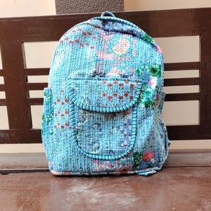 May include: A quilted backpack with a light blue background and a floral pattern in shades of red, pink, and green. The backpack has a front pocket with a curved flap and a blue trim. The bag is sitting on a brown wooden surface.
