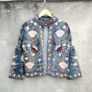 May include: A teal velvet jacket with intricate floral embroidery in shades of pink, brown, and cream. The jacket features a high collar and a vertical striped panel down the center. The embroidery creates a detailed, textured design.