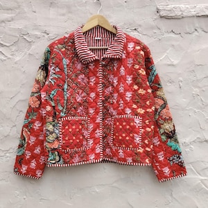 May include: A red quilted jacket with a floral design and two front pockets. The jacket has a striped collar and cuffs. The floral design includes pink, yellow, and green accents. The jacket is hanging on a wooden hanger.