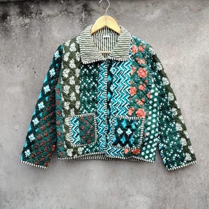 Peut inclure: Veste matelassée avec un motif patchwork. La veste présente une variété de motifs, notamment floraux, géométriques et à pois, dans des tons de vert, de sarcelle et de rouge. Le col est rayé et la veste est zippée.