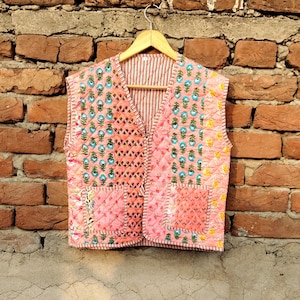 May include: A quilted pink vest with floral and geometric patterns, hanging on a wooden hanger against a brick wall. The vest features a V-neck, two pockets, and a striped lining. The patterns include blue and green floral motifs.
