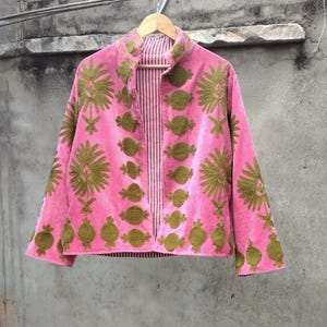 Peut inclure: Veste en velours rose ornée de motifs floraux et géométriques brodés vert olive. La veste a un col mandarin et une doublure rayée. La veste est suspendue à un cintre en bois.