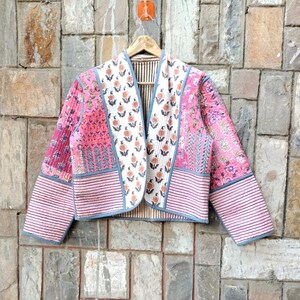 May include: A quilted jacket with a floral and striped pattern in shades of pink, white, and blue. The jacket is hanging on a wooden hanger against a stone wall. The jacket has a collarless design and long sleeves.