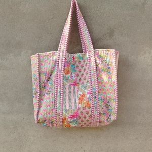 May include: A pink and floral patterned tote bag with matching handles. The bag is made of quilted fabric and features a variety of colorful floral designs. The bag is hanging against a grey wall.