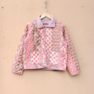 May include: A quilted jacket featuring a patchwork design with various pink floral patterns and geometric prints. The garment has a collared neckline, long sleeves, two front patch pockets, and a striped trim along the edges and collar.
