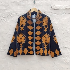 Op de afbeelding: Een donkerblauwe jas met ingewikkelde gouden borduursels. De jas heeft een opstaande kraag en een wit gestreepte rits. De borduursels bevatten bloemen- en cirkelpatronen, wat een gedetailleerd ontwerp creëert.