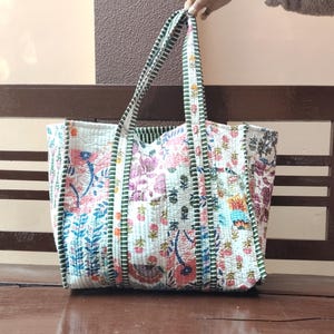 May include: A large, quilted tote bag featuring floral and paisley patterns in pink, blue, and green on a white backdrop. The bag has matching handles and trim, and is placed on a wooden bench.