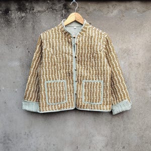 May include: A brown and white patterned jacket with a mandarin collar and two front pockets. The jacket is buttoned up and has light blue trim on the pockets and cuffs. The jacket is hanging on a wooden hanger.