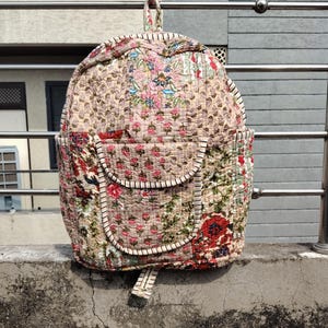 Puede incluir: Mochila acolchada con estampado floral en tonos rosa, rojo, verde y beige. La mochila tiene una parte superior redondeada, dos bolsillos delanteros y ribetes blancos. Cuenta con un asa superior y correas ajustables.