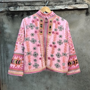 May include: A pink embroidered jacket with a mandarin collar and floral detailing. The jacket features gray, pink, and orange floral embroidery, with decorative patterns on the sleeves and collar. The jacket is hanging on a wooden hanger.