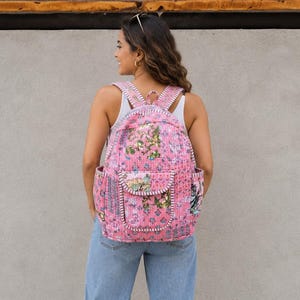 Könnte beinhalten: Ein rosafarbener, gesteppter Rucksack mit einem Blumenmuster in Grün-, Blau- und Weißtönen. Die Tasche verfügt über eine Vordertasche mit Klappe, Seitentaschen und verstellbare Schultergurte mit weißen Ziernähten an den Kanten.