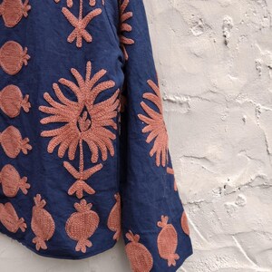May include: A navy blue garment with intricate coral-colored embroidered floral and fruit patterns. The embroidery is detailed, creating a textured effect on the fabric. The design is visible on the sleeve and body of the garment.