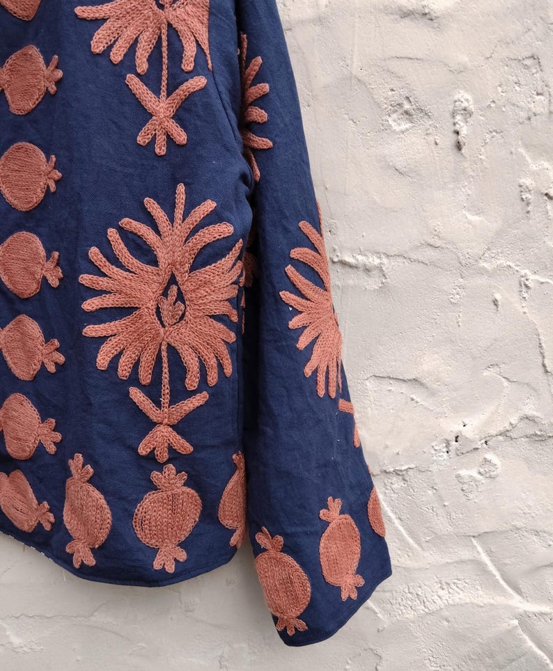 May include: A navy blue garment with intricate coral-colored embroidered floral and fruit patterns. The embroidery is detailed, creating a textured effect on the fabric. The design is visible on the sleeve and body of the garment.