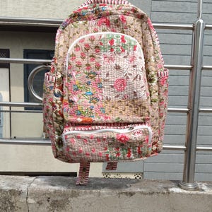 Puede incluir: Mochila acolchada con estampado floral en tonos rosas, verdes y beige. La mochila tiene un compartimento principal grande, un bolsillo frontal y bolsillos laterales. Cuenta con cremalleras blancas y un asa a rayas.