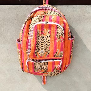 Puede incluir: Una mochila acolchada con un vibrante patrón de rayas verticales naranjas y rosas con un estampado de tigre repetitivo. La bolsa incluye un compartimento principal grande, un bolsillo frontal más pequeño y dos bolsillos laterales, todos asegurados con cremalleras blancas.