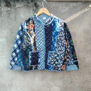 May include: A patchwork jacket with a variety of floral and geometric patterns in shades of blue, teal, and navy. The jacket has two front pockets and is lined with a striped fabric. It is hanging on a wooden hanger.