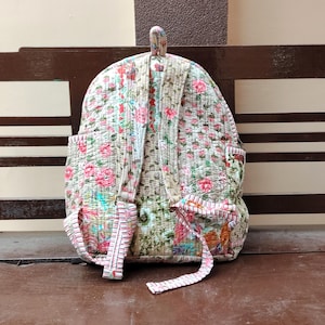 Puede incluir: Mochila acolchada con un estampado floral en rosa, rojo y verde sobre un fondo crema. La mochila tiene dos bolsillos laterales y ribetes rosas. Las correas y el asa también tienen un estampado floral.