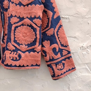 以下が含まれることがあります： ネイビーブルーのジャケットのクローズアップ。複雑なサンゴ色の刺繍が施されています。刺繍は花柄と幾何学模様で、テクスチャと詳細なデザインを作り出しています。ジャケットの袖が見え、手の込んだステッチが施されています。