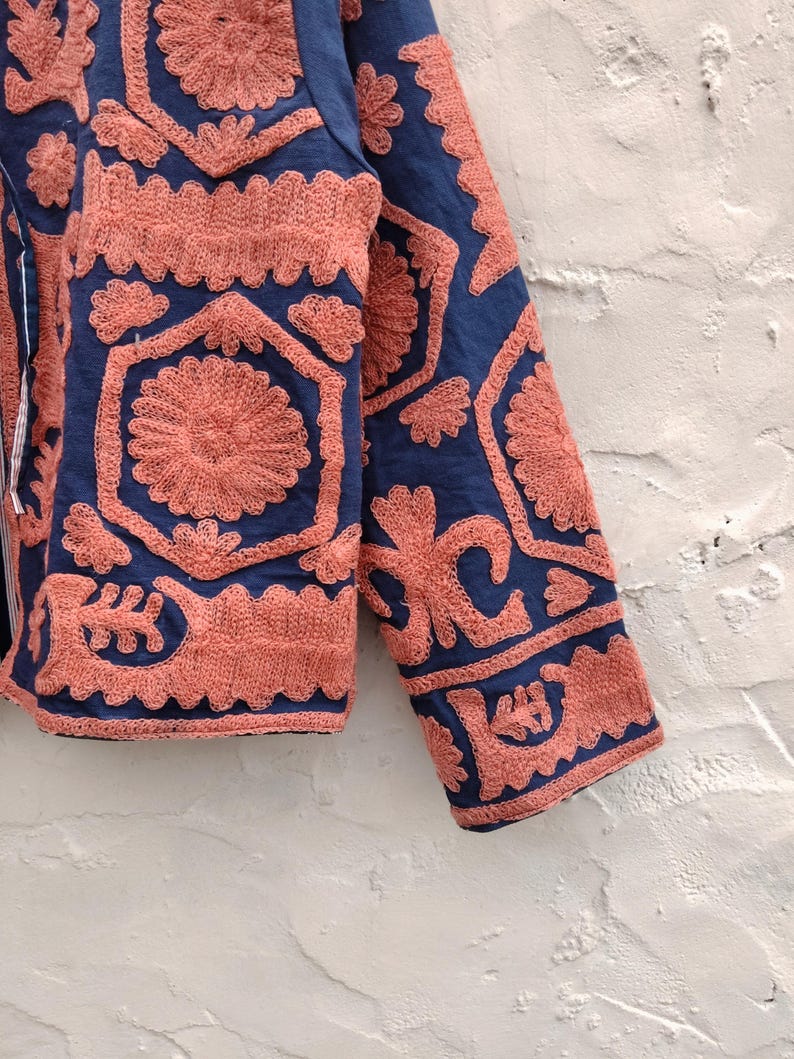 以下が含まれることがあります： ネイビーブルーのジャケットのクローズアップ。複雑なサンゴ色の刺繍が施されています。刺繍は花柄と幾何学模様で、テクスチャと詳細なデザインを作り出しています。ジャケットの袖が見え、手の込んだステッチが施されています。