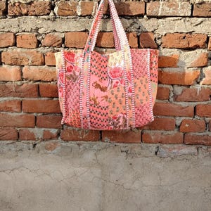 May include: A pink tote bag with floral and animal print patterns hangs against a brick wall. The bag features a patchwork design with various shades of pink, red, and orange. The handles are made of the same patterned fabric.