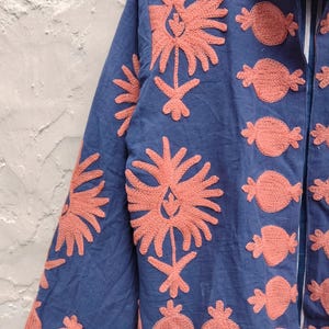 May include: A blue jacket with coral-colored embroidered floral and fruit designs. The jacket features long sleeves and a collared neckline. The embroidery is detailed and covers the front, sleeves, and collar of the jacket.