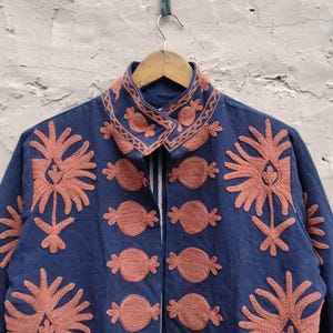 May include: A navy blue jacket with intricate coral-colored embroidery. The jacket features a collared neckline and a series of floral and abstract designs. The embroidery is detailed and covers the front and collar of the jacket.