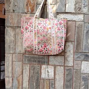 May include: A floral tote bag with a quilted design. The bag features a pattern of pink and red flowers with green leaves on a cream background. The handles and side panels have a striped pattern. The bag is hanging against a stone wall.