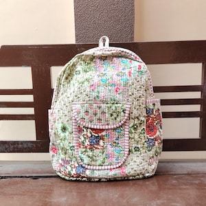 Puede incluir: Una mochila acolchada con un estampado floral en tonos verdes, rosas y azules. La mochila tiene dos bolsillos delanteros con ribete rosa y un lazo para colgar. La bolsa está sobre un banco de madera.