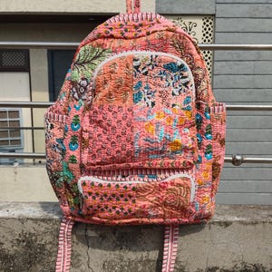 May include: A patchwork backpack in pink, orange, and blue, with floral and geometric patterns. It has multiple pockets, white zips, and adjustable straps. The bag is made of quilted fabric.