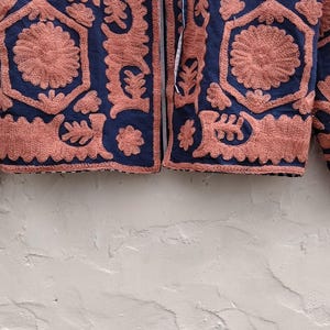 以下が含まれることがあります： ネイビーブルーのジャケット。サンゴ色の刺繍が施されており、花柄や幾何学模様が特徴的で、テクスチャードデザインを作り出しています。 ジャケットはジッパーフロントと長袖で、色とテクスチャの組み合わせを表現しています。