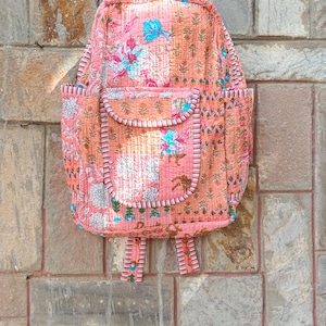 May include: A quilted backpack in shades of pink and peach, adorned with floral patterns in white and blue. The backpack features a front pocket, side pockets, and adjustable straps. The design includes a top handle for easy carrying.