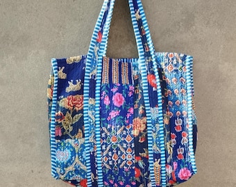 May include: A blue tote bag with floral and animal print patchwork. The bag has blue and white striped handles and trim. The bag is made of quilted fabric and is hanging against a grey wall.