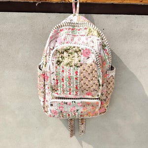 Op de afbeelding: Een gewatteerde rugzak met een patchworkontwerp met diverse bloemen- en botanische patronen in tinten roze, groen, beige en wit. De tas bevat een groot hoofdvak, een voorvak met ritssluiting en zijvakken met een witte afwerking.