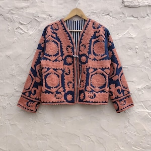 以下が含まれることがあります： ネイビーブルーのジャケット。複雑なコーラル色の刺繍が施されています。 ジャケットは花柄と幾何学模様で、ストライプの裏地と紐留めが付いています。 木のハンガーに掛けられ、質感のある壁に掛けられています。