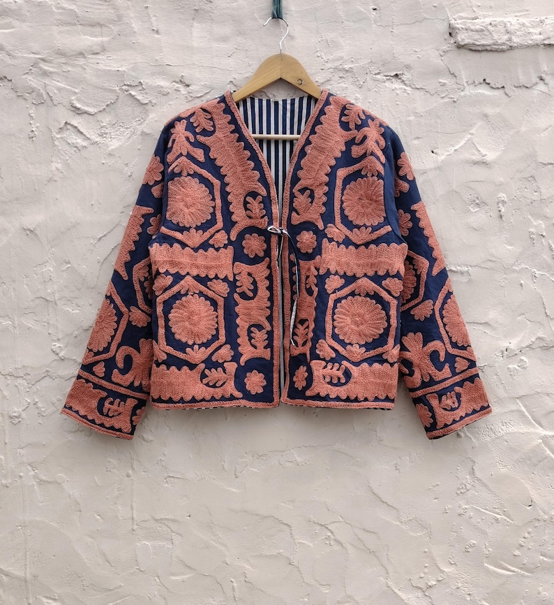 以下が含まれることがあります： ネイビーブルーのジャケット。複雑なコーラル色の刺繍が施されています。 ジャケットは花柄と幾何学模様で、ストライプの裏地と紐留めが付いています。 木のハンガーに掛けられ、質感のある壁に掛けられています。