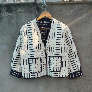 Pode incluir: Casaco acolchoado creme com um padrão geométrico preto. O casaco apresenta decote em V, dois bolsos frontais e botões de madeira. Os punhos e a gola são adornados com um tecido azul escuro. O casaco está pendurado num cabide de madeira.