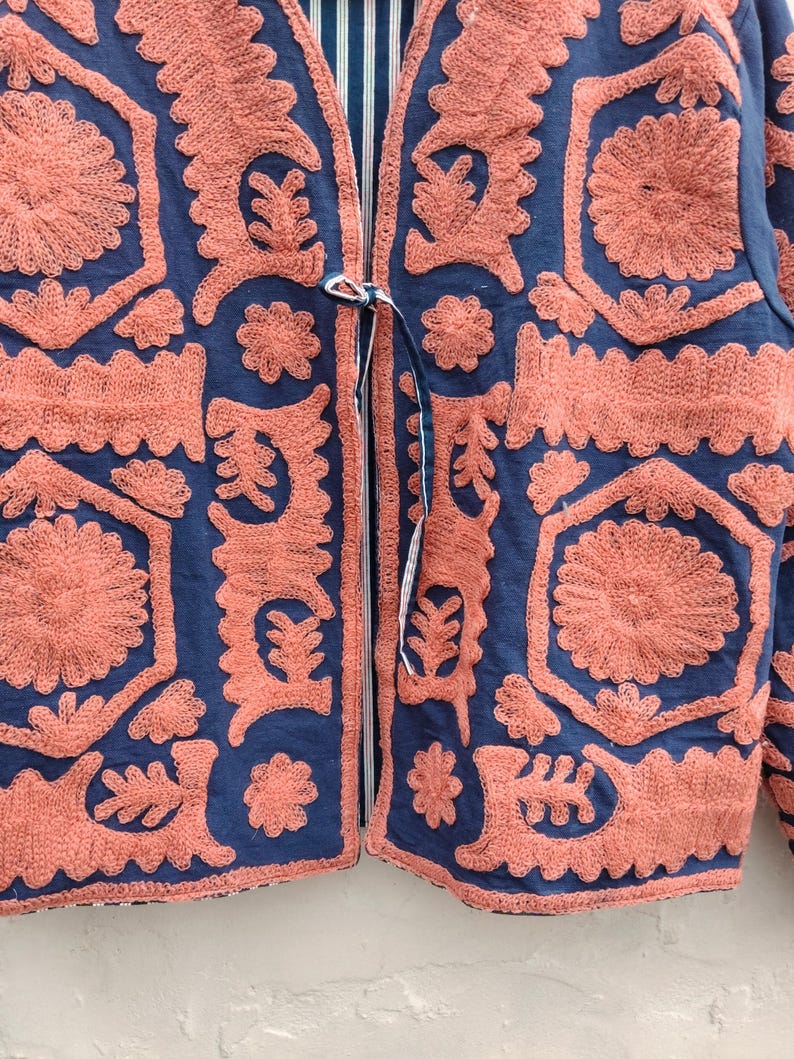 以下が含まれることがあります： ネイビーブルーのジャケット。サンゴ色の刺繍が施されており、結び紐で留めるデザイン。裏地はストライプ柄。刺繍は花柄や幾何学模様で、細部までこだわったテクスチャードデザイン。